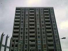 Eleven storeys of a gray residential tower, with a spiked fence and a lamp in the foreground.