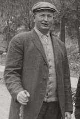 A man in a cap and jacket walks on a dirt road carrying a walking stick in his right hand.