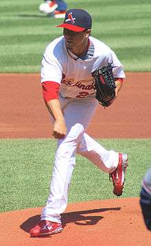 A man wearing a white baseball uniform and a navy-blue baseball cap with a red brim and a bat and cardinal bird on the face throws a baseball (unseen) from a dirt mound