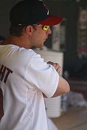 A man in a white baseball jersey, navy blue baseball cap with a red brim, and orange mirrored sunglasses stands with his arms crossed.