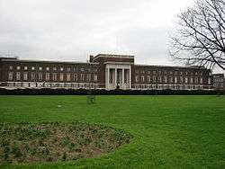 Very wide flat-roofed mulberry brick building with three rows of widows, portico is Portland stone with four columns