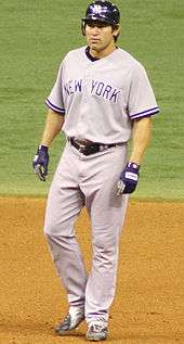 A man in a gray baseball uniform with "NEW YORK" on the chest and a dark batting helmet stands in the infield dirt of a baseball diamond.