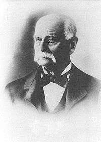 Head shot of an older man with white hair and moustache, dressed in tuxedo and bow tie.