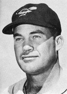 A man wearing a white baseball jersey and a black baseball cap with a bird on the face