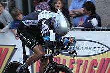 A road racing cyclist, wearing a black and white skinsuit with blue trim and an aerodynamic helmet, sits crouched very low on his bicycle. Spectators watch from the roadside.