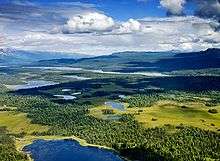 Aerial photograph of several lakes, interspersed with conifer forests and meadows, with tall mountains in the distance and clouds and blue sky overhead. The clouds are casting shadows over the forests and lakes.