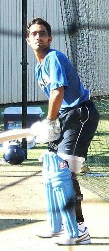 Brown-skinned young man, not clean shaven, wears a sky blue shirt with the words "SAHARA". He is wearing white sports shoes, navy shorts and black stockings, is wearing light blue cricket pads on his legs, white gloves, is holding a bat and is capless, in batting stance on a cricket pitch.