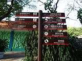 A sign labeled with directions pointing to a tourist centre, a clinic, a park exit, an animals baby farm, a peacock garden, a zoology museum, an Amusement World, and a wetland garden.