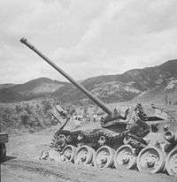 A damaged main battle tank, lying to one side of the road with its main gun traversed and its tracks blown off