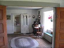 Room with oval braided rug on wood floor, door in opposite wall, small organ in corner, window at right
