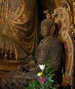 Three-quarter view of cross-legged seated statue placed on a throne. Both arms rest on his legs. The hair is sculpted with a top knot, which supports a sacred casket.