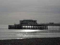 The end of a pier under cloudy skies, illuminated by sunlight reflecting off a calm sea.  There is a small stretch of rocky beach in the foreground.