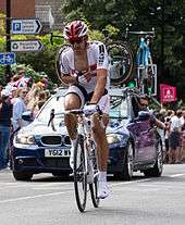 A cyclist riding a bike while wearing a white uniform and holding his right arm to his chest.