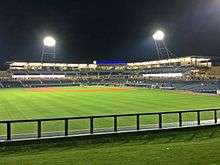 A nighttime view of the gray concrete concourse, blue seats, and green playing field illuminated by two lighting stanchions on the concourse after game