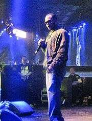 A black man with short black hair. He wears dark sunglasses of an unrecognizable color, a brown jacket, and blue jeans. Two men with brown hair stand far behind him, wearing black shirts with unrecognizable yellow text.