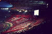 A full stadium, with fans holding up white and red cards to make a giant mosaic.