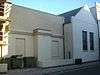 Side view of a two-part, plain-walled building in shadow. The nearer section is flat-roofed, painted cream and has two blank windows with a wide pillar between them.  Rubbish bins and scaffolding are to the left.  The far section is white, has a pointed roof whose end resembles a pediment, and has two identical red-framed windows.