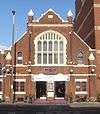 Front view of a squat, red-brick building with a steep pointed roof and prominent stone dressings. There are four identical spirelets: two ending about halfway up the building and two level with the top of the roof.  A pointed-arched window, split by mullions into ten panes, dominates the façade.  Below it are two entrance doors and the word "ten" in purple lower-case letters.