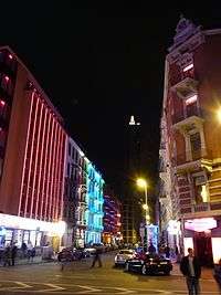 A city street at night, with many colourfully lit buildings. On the left is a large, red tower.
