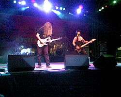 A medium sized rockscene lit in many colors. In the left stands a longhaired guitarist on the right a bass player wearing an armystyle west and helmet. In the background are two drumkits with a drummer.