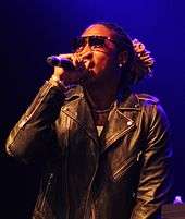 A portrait of an African-American man with dreadlocks, performing on stage, wearing a jacket and sunglasses.