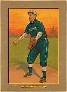 A man in a navy blue baseball uniform with white cap and undershirt long sleeves stands on a baseball field with his arms spread apart awaiting a ball to be thrown.