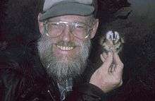 A smiling, bearded man holds a small bird near his left ear with his right hand.