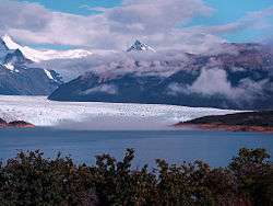 A glacier flows down to a bright blue lake surrounded by snow-capped mountains