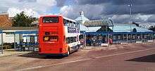 A bus concourse with bus station building to the left. The bus station has blue cladding and glass and a lantern feature on the roof with a clock and windvane