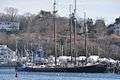 Adventure is a long black ship with three masts. She sits relatively low to the water, anchored at a berth in front of other smaller ships, and land rising in the background.