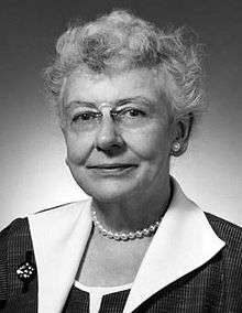 a black-and-white head-and-shoulders portrait of a smiling grey-haired woman wearing a smart dress, a pearl necklace and earring, a small broach and rimless pince-nez spectacles.