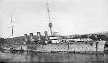 A four funnelled warship lies at anchor in a harbour.