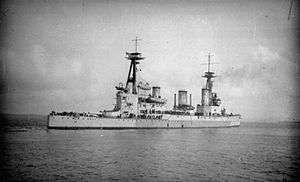 Right rear oblique shot of a large gray warship with two masts, three funnels, and two gun turrets visible. A number of people are visible on her decks.