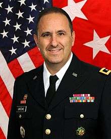 A photograph of a grinning man in a United States Army uniform. Behind the man are two separate flags that comprise the background, one being the national flag of the United States of America and the other being the personal standard of a United States Army major general.