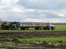 A farm field with a white tractor towing a long wagon with wood-plank walls used to collect vegetables. A farmer is standing on the back of the wagon, adjacent to a harvesting machine which uproots carrots from the field and sends them along a slanted conveyor belt to the farmer. In the foreground is a brown-coated dog.