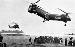 Black and white image of a large group of refugees in flood waters below two helicopters.