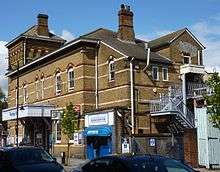 A brick building with decorative brickwork; a tower can be seen to the left and a stairway to the right. The main entrance is at the centre of the building; a taxi office is to the entrance's right.