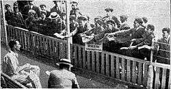 A cricketer sitting on a chair with a large group of boys watching, holding paper for him to autograph