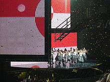 Far away image of a group of people on stage, wearing white vests and long patterned skirts