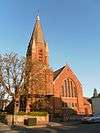 A dark red church dominated by a tower on the left. This has stone spirelets at each corner and a central slate-coloured spire, and is partly obscured by trees.  There are five lancet windows in a slight recess in the wall to the right.