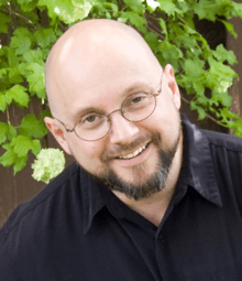 Picture of Howard Tayler wearing a medium blue denim button-up collared shirt. He is looking over the top of his glasses while grinning at the camera.