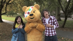 A young girl, a person dressed as a large smiling, yellow teddy bear and a young man stand in a grassy park, with their hands over their hearts. Their mouths are open, as if singing. The girl is wearing a blue jacket and a yellow scarf. The man is wearing a checked shirt and a white T-shirt. The teddy bear outfit has a polka dot bandana stitched over the bear's right eye.