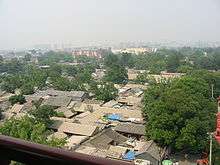 A view down to the gabled roofs of single-story houses with some interspersed trees in the foreground. In the background, slightly obscured by smog, are taller, more modern buildings