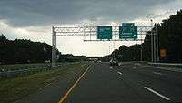 A four lane freeway at an interchange in a wooded area with two green signs over the road. The left one reads Interstate 195 west Trenton and the right one reads exit 21 County Route 527 County Route 526 Jackson Siloam upper right arrow