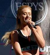 With slicked back blonde hair, a woman holds a microphone.