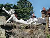 Colour photograph of an individual female statue with short tousled hair lying on a rock pedestal. Supporting herself on her left arm, her head is up and her right arm raised