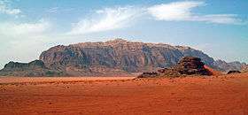 A wide rocky tan-colored ridge, reaching a high point in the middle, over a desert of reddish sand with a smaller prominence in the foreground