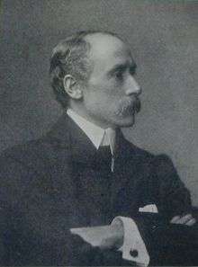 Black-and-white photographic profile portrait of a man in his forties, with a strong nose, moustache, receding hair, in a suit with his arms folded.