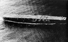 An aerial view of an aircraft carrier at sea. Her small island is on the left side and only three aircraft are visible on her flight deck at the bow.