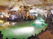 Brightly illuminated cave traversed by a bright emerald colored subterranean body of water encircling a variety of speleothems in the background